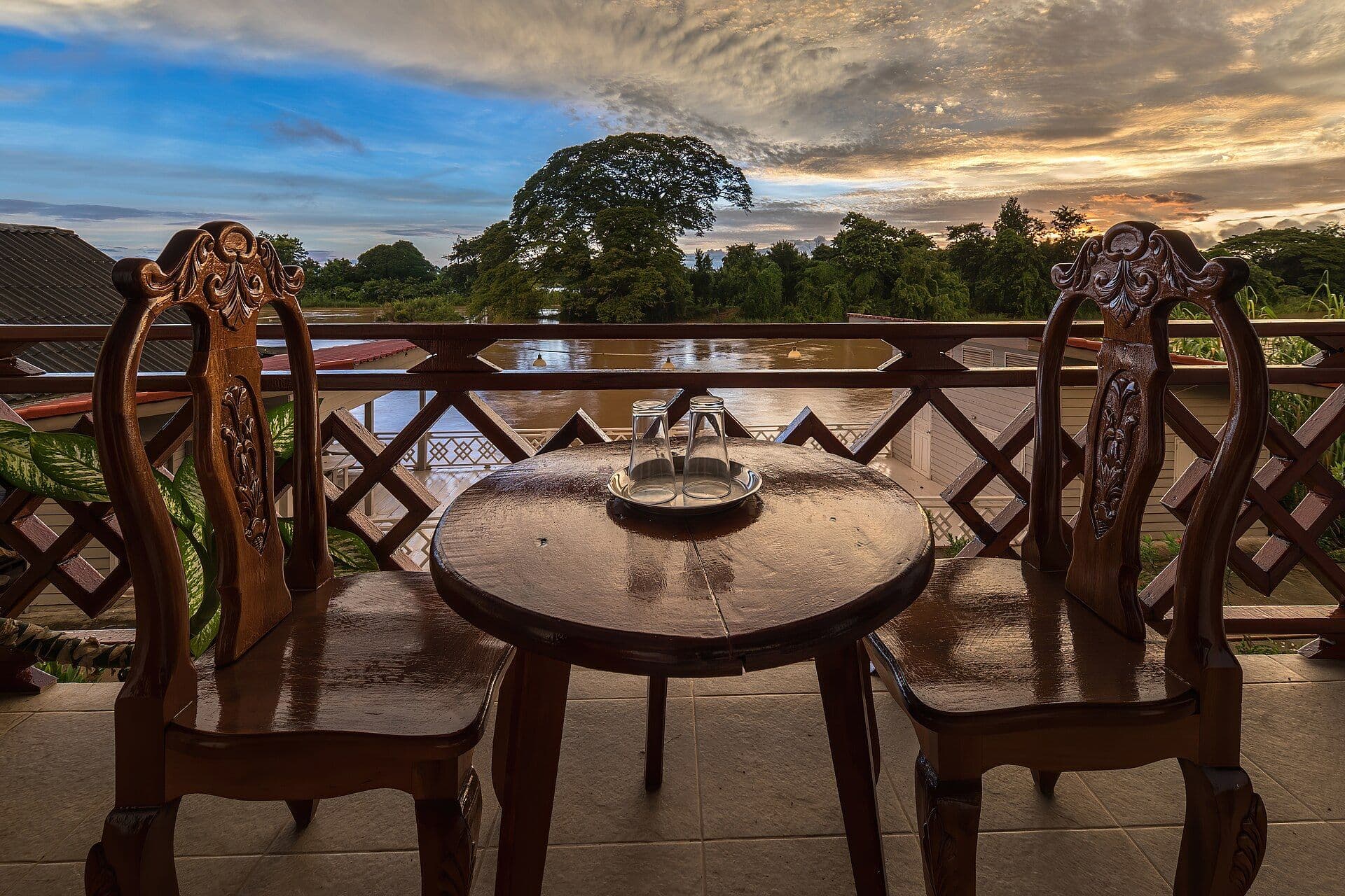 A round wooden table with two drinking glasses set between two carved wooden chairs on a balcony overlooking a river at sunrise.